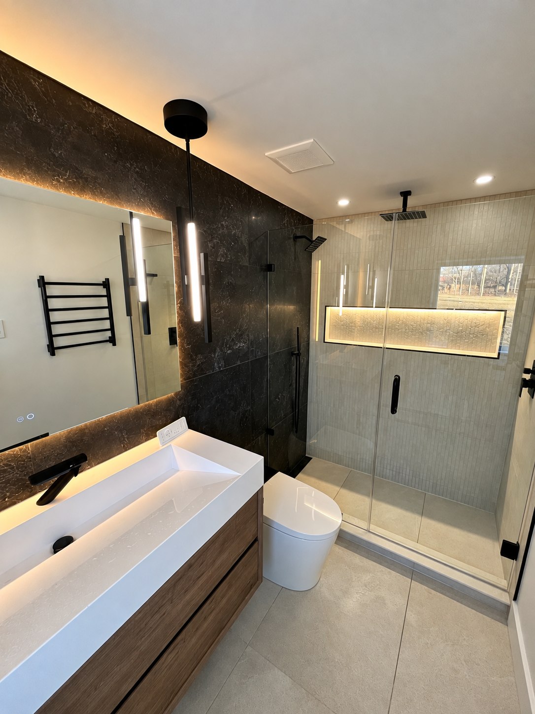 Contemporary bathroom with a wood vanity, illuminated mirror, dark stone accent wall, frameless glass shower, and backlit niche.