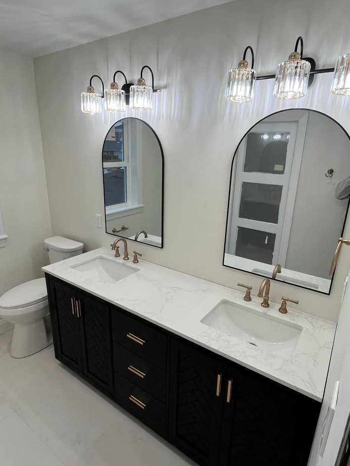 Black double vanity with a white stone top, brass hardware, arched mirrors, and crystal-style sconces.