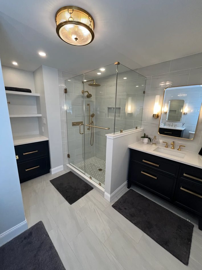 Bathroom with a dark vanity, gold hardware, glass shower, light tile, built-in shelving, and layered lighting.