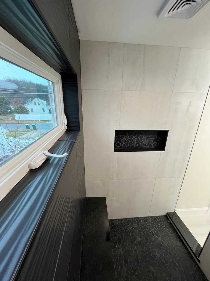Walk-in shower with black pebble floor, oversized white wall tile, black-framed window, and recessed black niche.