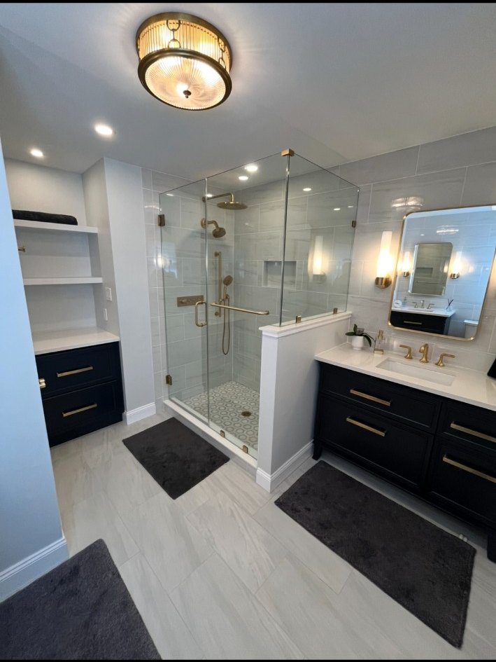 Wide bathroom view with a brass-trimmed frameless shower, patterned shower floor, dark vanity, built-in shelving, and custom lighting.