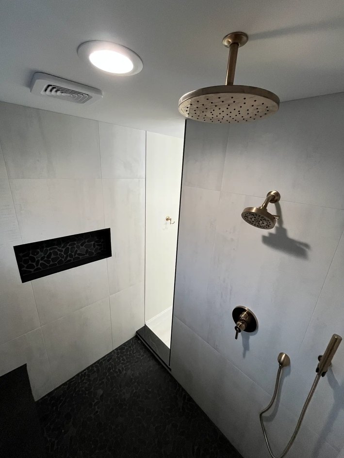 Spacious bathroom with a glass corner shower, blue-gray tile, warm metal fixtures, dark vanity, and built-in shelving.