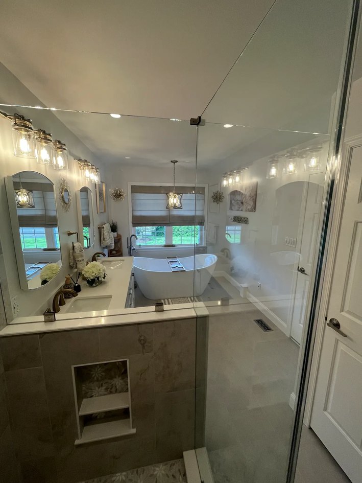 View through a clear glass shower divider toward a freestanding tub, vanity, mirrors, and pendant light.
