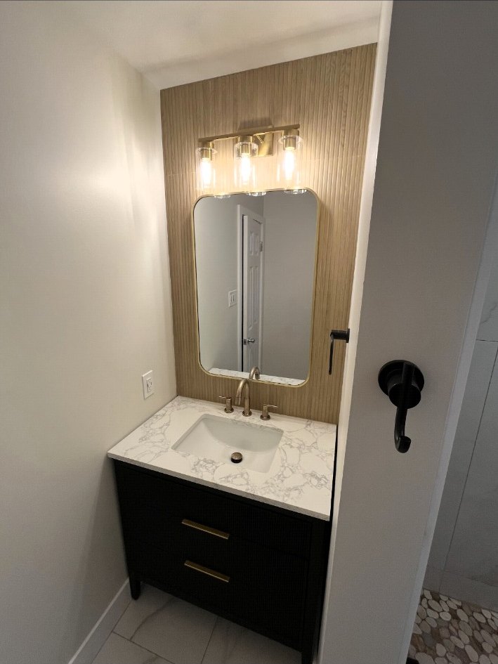 Bathroom vanity with a veined stone countertop, brass faucet, black cabinetry, wood-slat wall, mirror, and glass sconces.