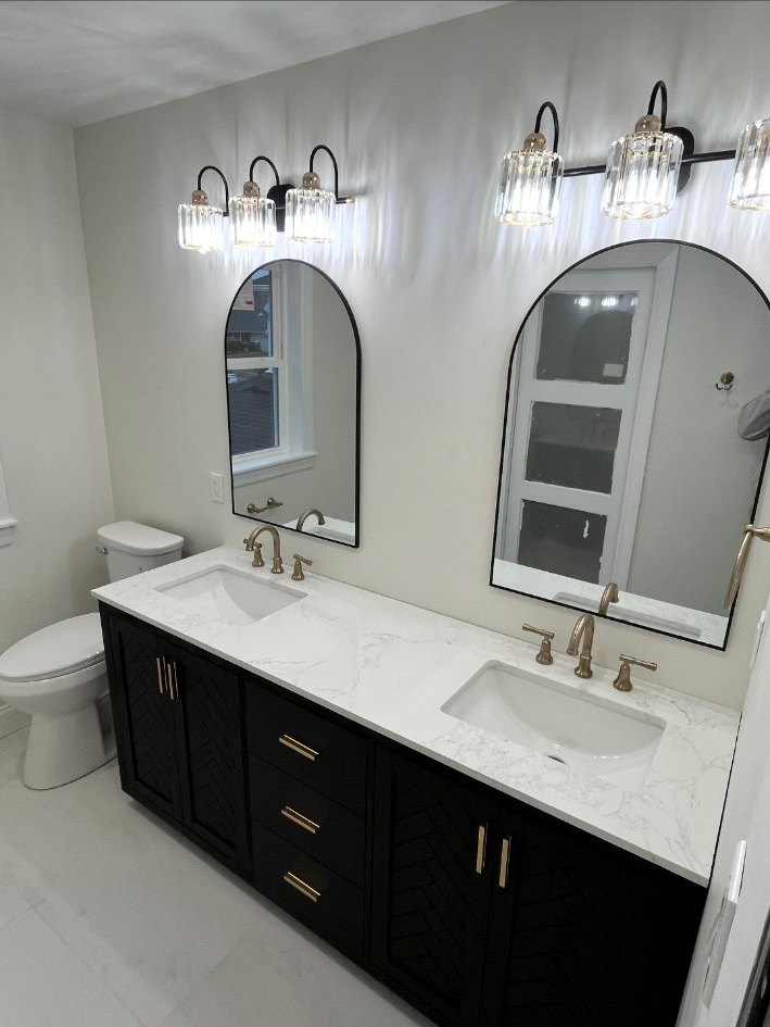 Dark double vanity with a white stone countertop, brass faucets, arched mirrors, and ribbed-glass sconces.