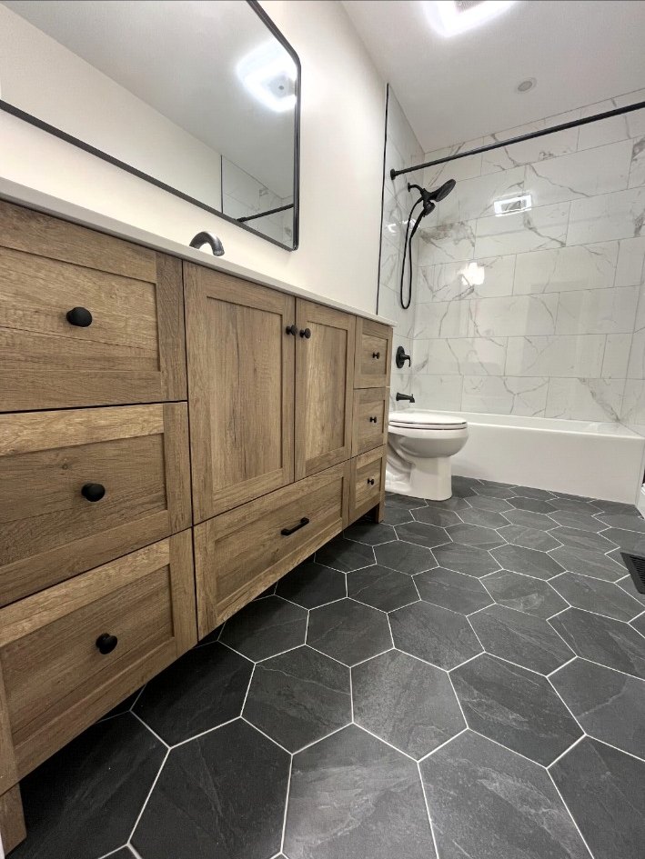 Low view across a wood vanity and dark hexagonal floor toward a white tub with stone-look tile surround.