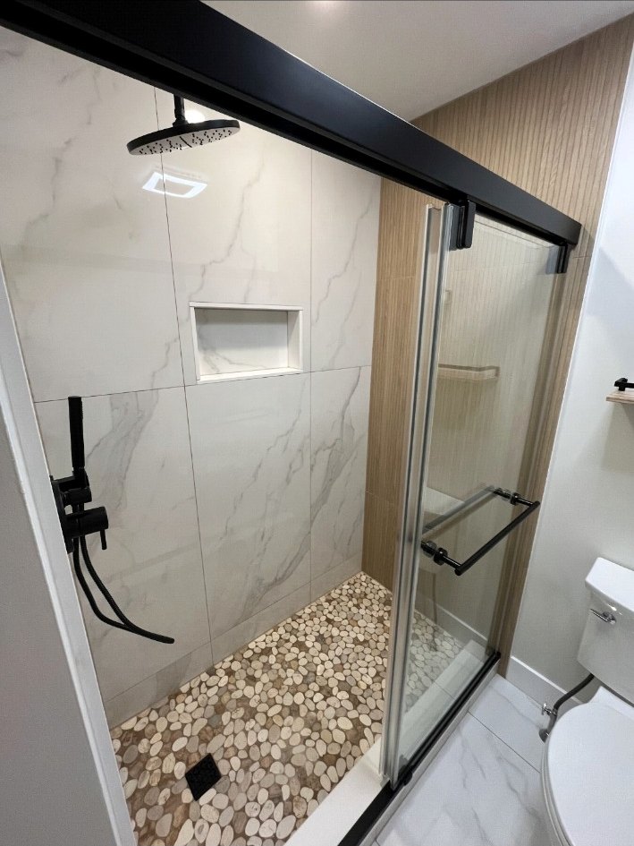 Frameless shower with large-format marble-look tile, black sliding hardware, rain head, niche, and pebble floor.