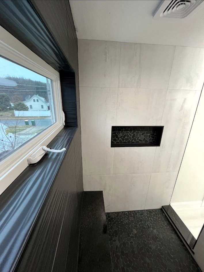 Large-format tiled shower with a dark pebble niche, matching shower floor, black bench, and window detail.