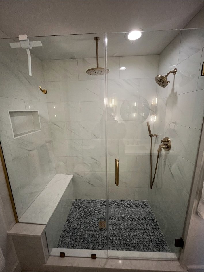 Frameless glass shower with brass hardware, ceiling rain head, wall shower, handheld fixture, bench, niche, and pebble floor.