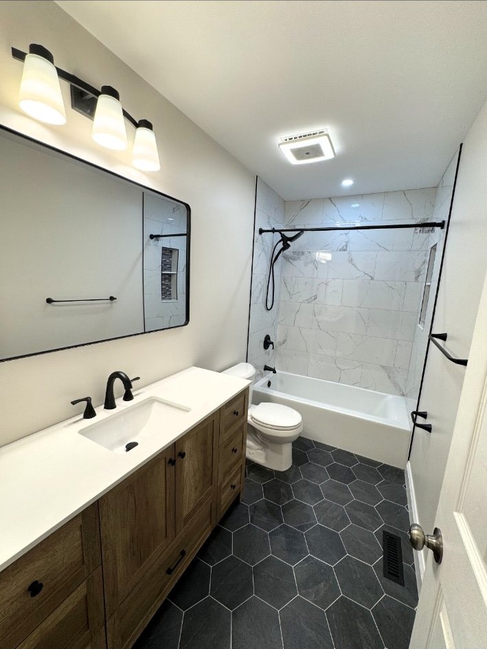 Full bathroom with a natural wood vanity, dark hex floor tile, black-framed mirror, and marble-look tiled tub surround.