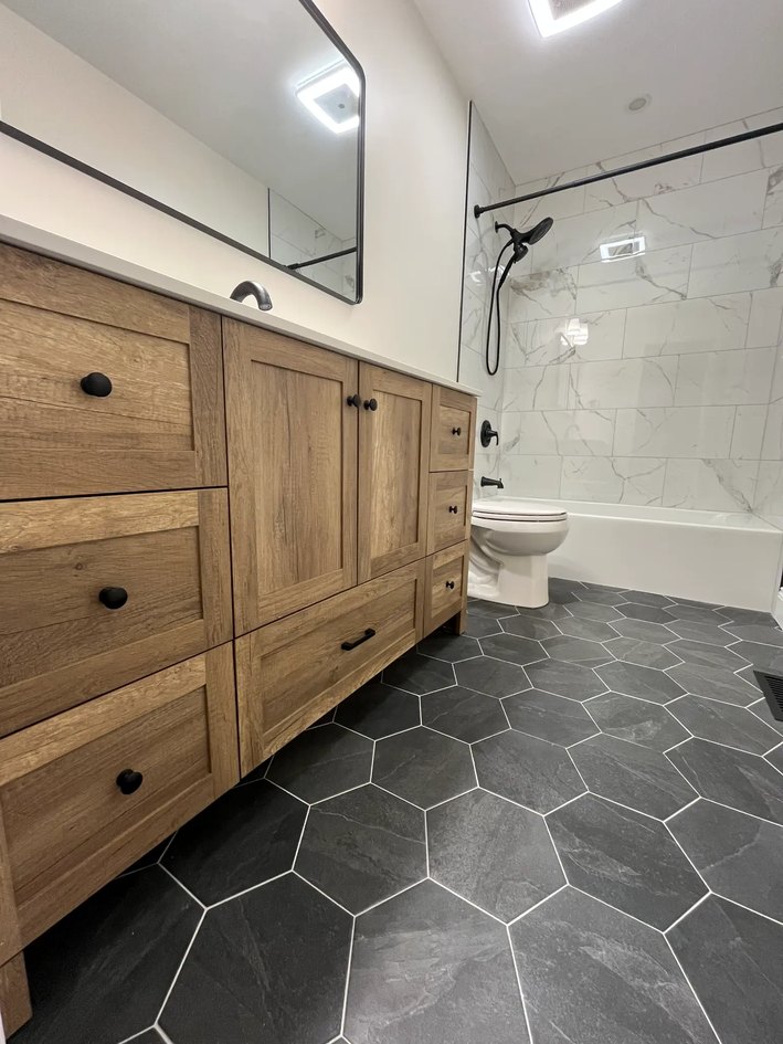 Oak vanity and dark hex floor leading toward a tiled tub surround.