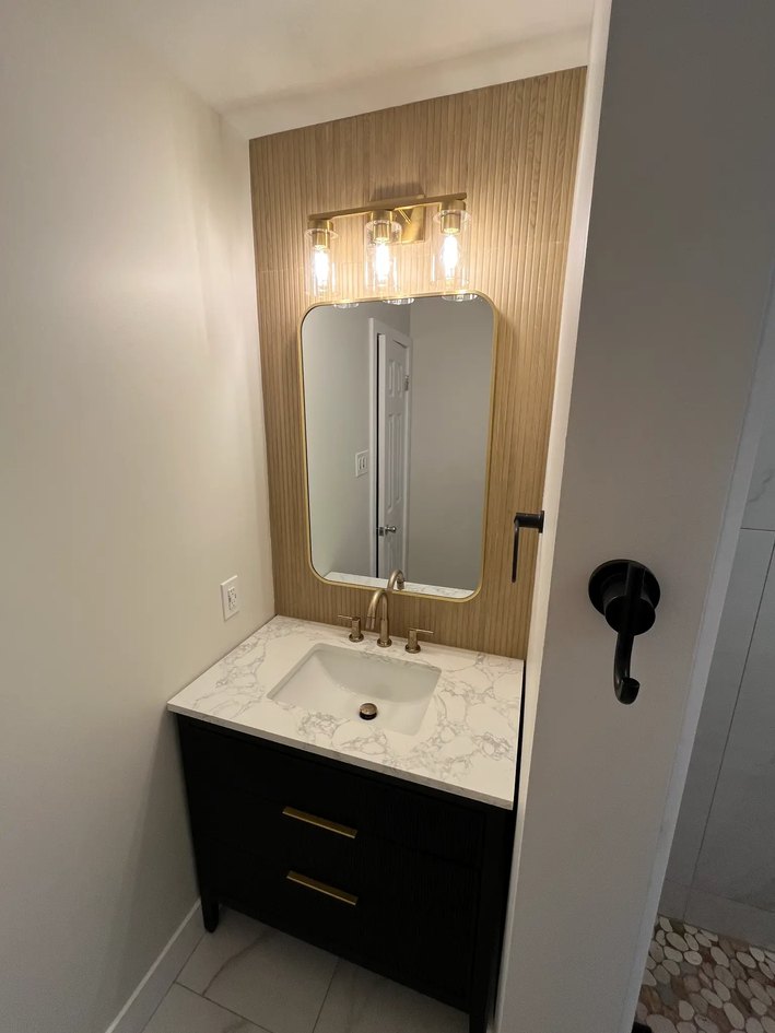 Compact black bathroom vanity with a veined white top, rounded mirror, warm slatted wood wall, and brass lighting.