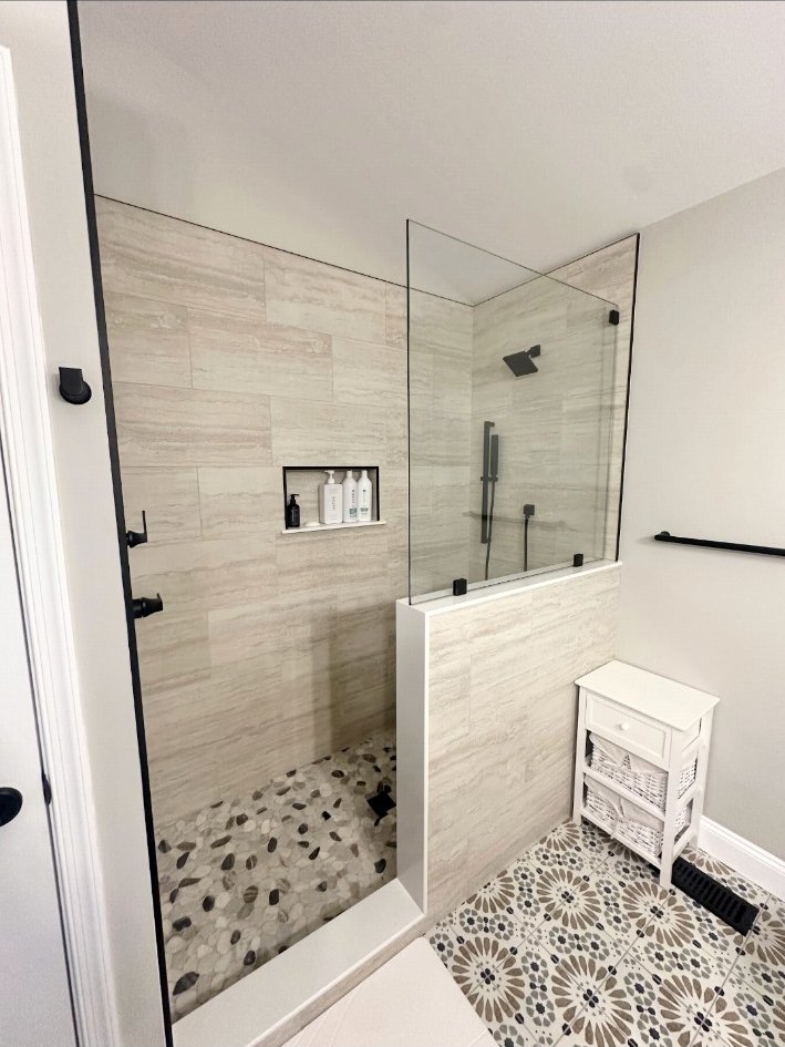Frameless walk-in shower with matte-black fixtures, a recessed niche, neutral wall tile, pebble floor, and patterned bathroom floor.