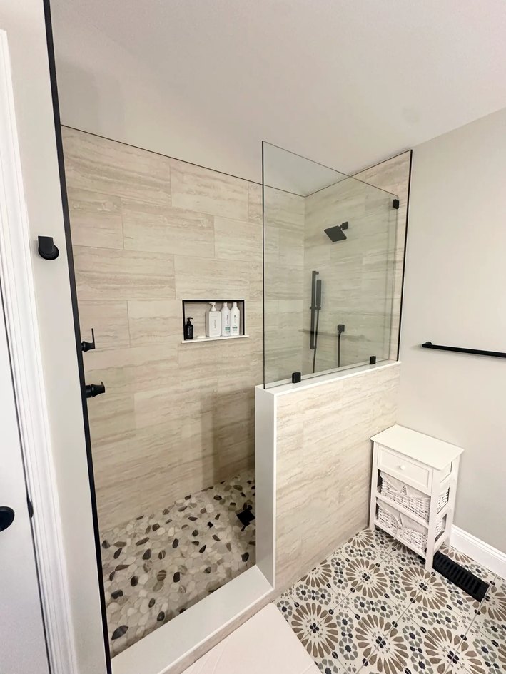 Glass-enclosed shower with white stone-look tile, pebble floor, bench, rain head, wall shower, and hand spray.